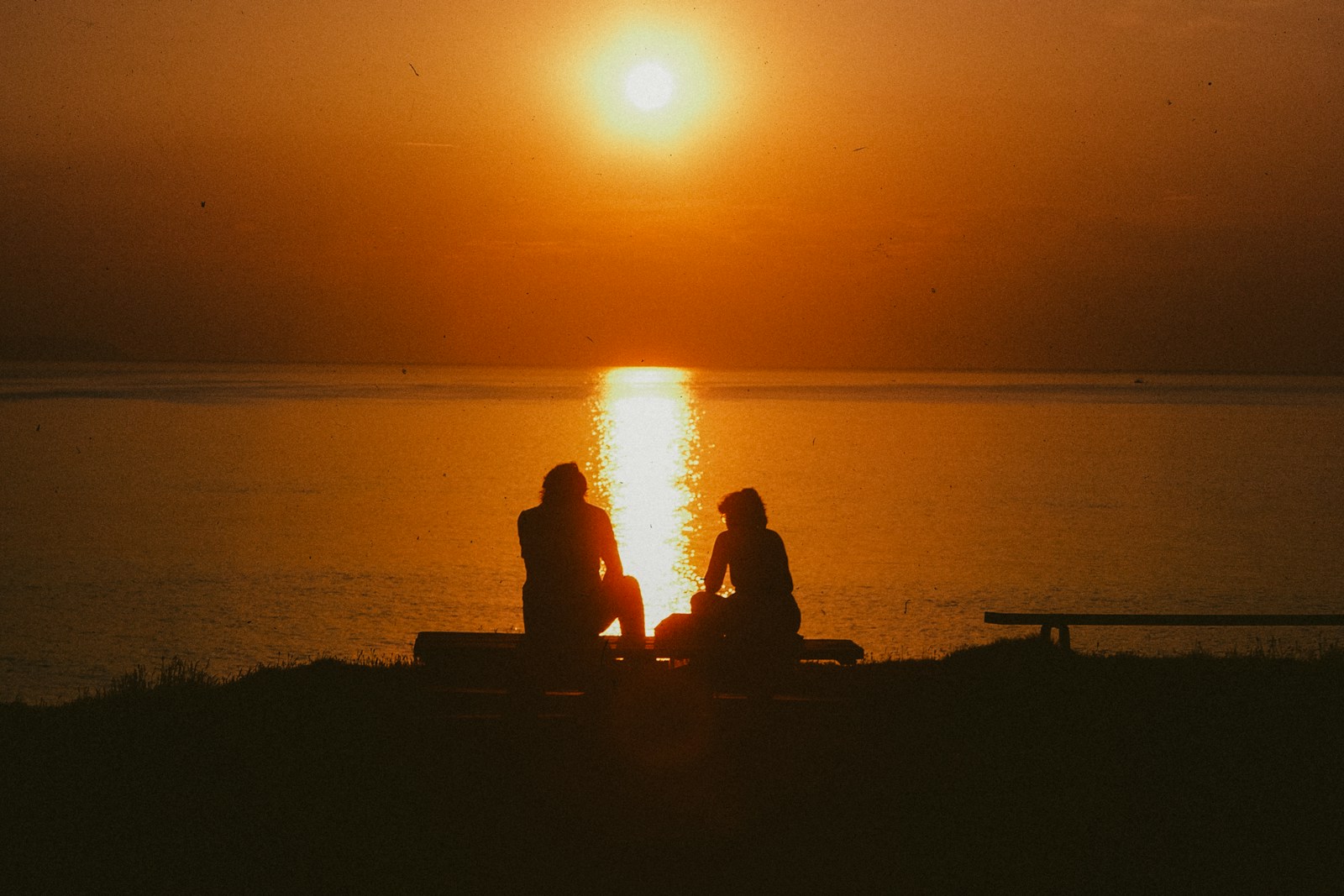 Two silhouettes sit by the ocean at sunset.